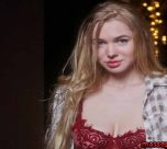Young woman with long blonde hair wearing a red lace bra and an open plaid shirt, standing in front of warm string lights in a dark backdrop.