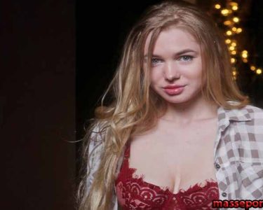Young woman with long blonde hair wearing a red lace bra and an open plaid shirt, standing in front of warm string lights in a dark backdrop.