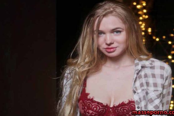 Young woman with long blonde hair wearing a red lace bra and an open plaid shirt, standing in front of warm string lights in a dark backdrop.