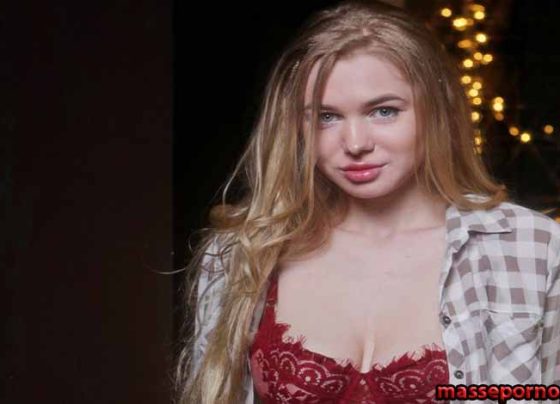 Young woman with long blonde hair wearing a red lace bra and an open plaid shirt, standing in front of warm string lights in a dark backdrop.