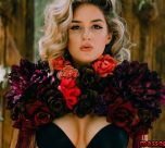 Glamorous portrait of a woman with blond curls wearing a black low-cut top, framed by a large red and burgundy floral collar.